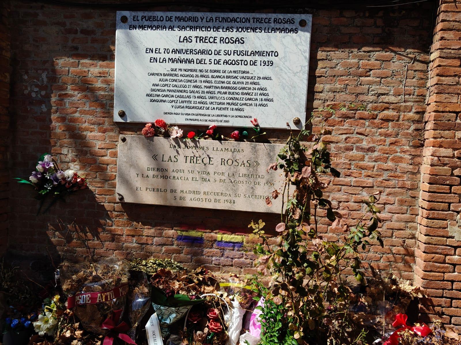 Placa conmemorativa sobre un muro de ladrillo.
Hay dos placas rectangulares, una de mármol blanco arriba y otra de piedra beige debajo, ambas sujetas con pernos metálicos.
Transcripción de la placa superior:
“EL PUEBLO DE MADRID Y LA FUNDACIÓN TRECE ROSAS
EN MEMORIA AL SACRIFICIO DE LAS JÓVENES LLAMADAS
LAS TRECE ROSAS
EN EL 70 ANIVERSARIO DE SU FUSILAMIENTO
EN LA MAÑANA DEL 5 DE AGOSTO DE 1939
QUE NI SU NOMBRE NI SU SUEÑO SE BORREN DE LA HISTORIA...
CARMEN BARRERO AGUADO 20 AÑOS.
BLANCA BRISAC VÁZQUEZ 29 AÑOS.
JOSÉFA CENDEJA 50 AÑOS.
ELENA GIL OLAYA 20 AÑOS.
ANA LÓPEZ GALLEGO 21 AÑOS.
JULIA CONESA CONDE 19 AÑOS.
VIRTUDES GONZÁLEZ GARCÍA 18 AÑOS.
DIONISIA MANZANERO SALAS 20 AÑOS.
PILAR BUENO IBARRA 22 AÑOS.
ADELAIDA GARCÍA CASILLAS 19 AÑOS.
VIRTUDES NÚÑEZ GARRIDO 18 AÑOS.
JOAQUINA LÓPEZ LAFUENTE 23 AÑOS.
CARMEN CUESTA GARCÍA 18 AÑOS.
LUISA RODRÍGUEZ DE LA FUENTE
FALLECIÓ EN PRISIÓN UNOS DÍAS ANTES DE LA EJECUCIÓN
EN MADRID A 5 DE AGOSTO DE 2009
LA DEMOCRACIA”
Transcripción de la placa inferior:
“LAS JÓVENES LLAMADAS
«LAS TRECE ROSAS»
DIERON AQUÍ SU VIDA POR LA LIBERTAD
Y LA DEMOCRACIA EL DÍA 5 DE AGOSTO DE 1939
EL PUEBLO DE MADRID RECUERDA SU SACRIFICIO
5 DE AGOSTO DE 1988”
Alrededor de las placas hay varios ramos de flores de distintos colores, algunos apoyados en la parte inferior del muro y otros colocados justo bajo las placas. También hay una planta de tallos largos y hojas marrones y verdes subiendo por la zona derecha de la placa inferior. En la parte baja del muro se ven más flores, coronas y envoltorios de plástico, como ofrendas.