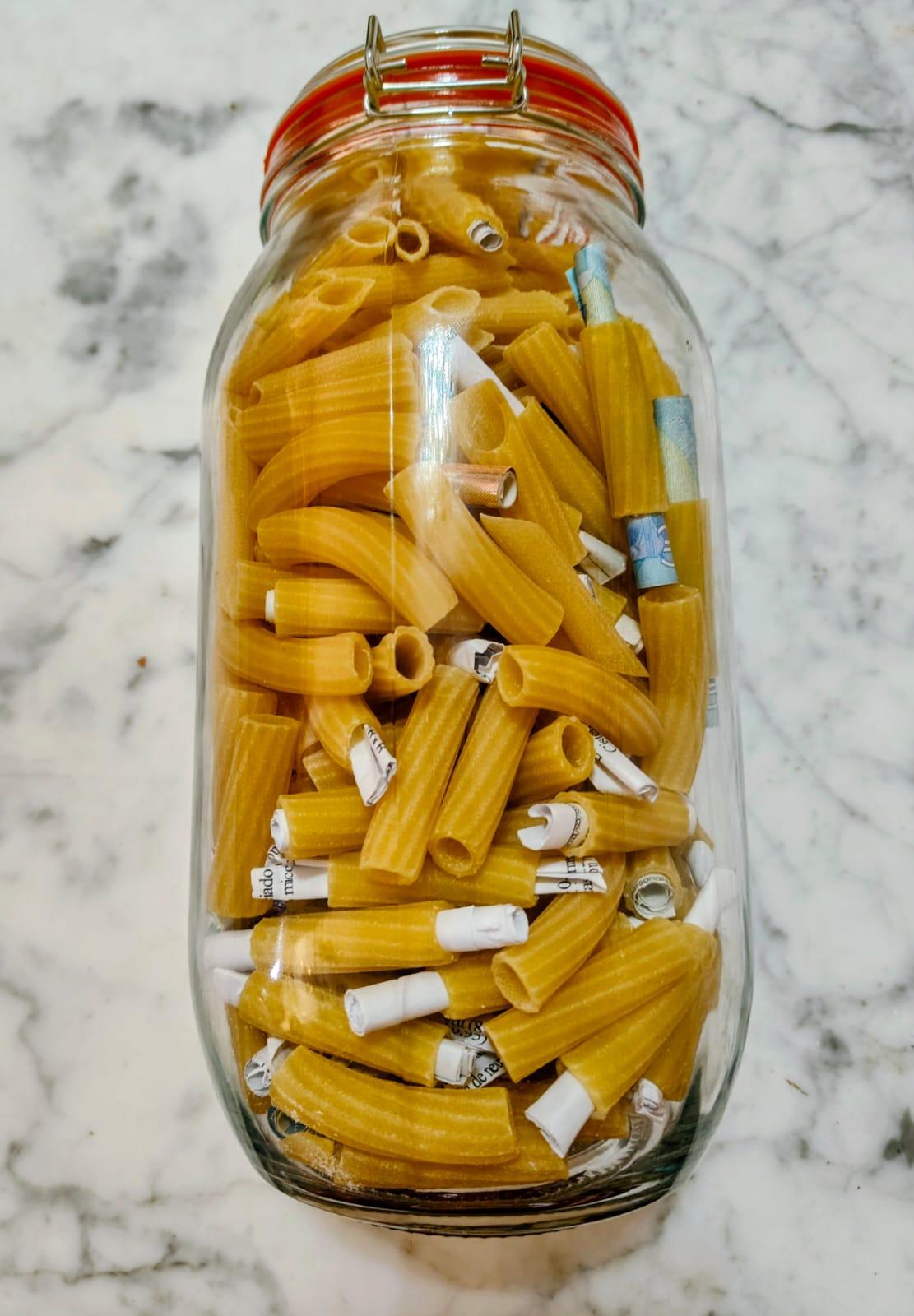 Un frasco grande de vidrio lleno de pasta seca tipo macarrón grueso.
La pasta es de color amarillo dorado y está colocada vertical y horizontalmente, ocupando casi todo el frasco.
Entre los macarrones hay muchos papelitos pequeños enrollados, de color blanco y algunos con partes azules o con letras impresas en negro.
El frasco tiene tapa hermética de metal con aro rojo.
El fondo es una superficie lisa de mármol claro con vetas grises.